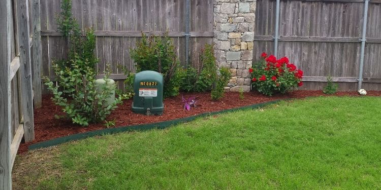 A garden bed in the corner of a fenced in back yard. It contains several small plants and fresh mulch.