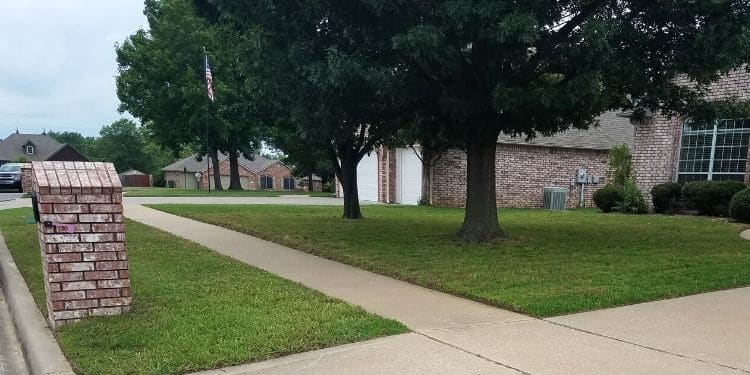 A front lawn serviced by Sprague's Kik'N Grass. The grass is green and uniformly mowed. There are two large trees in the foreground and nicey pruned shrubs in the background.